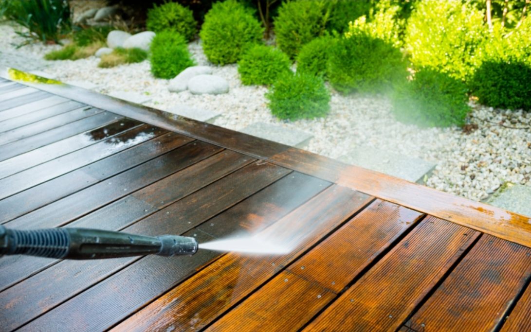 Un nettoyeur haute pression nettoie une terrasse en bois, entourée de verdure.